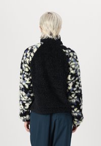 Fleece bunda s černým texturovaným zadním dílem, barevně vzorovanými rukávy v modré, černé a krémové barvě a s vysokým límcem.