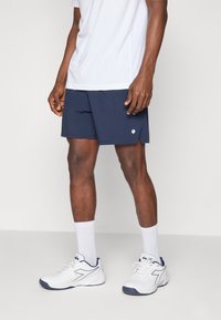 Bermudas atléticas azul-marinho com uma textura suave e fendas laterais, combinadas com uma t-shirt branca que afasta a humidade e sapatilhas brancas com detalhes azul-marinho.