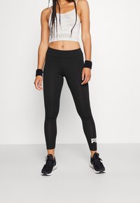 Kobieta ubrana w czarne legginsy Puma, białą krótką bluzkę na ramiączkach, czarne opaski na nadgarstki i czarne buty sportowe, stojąca na białym tle.