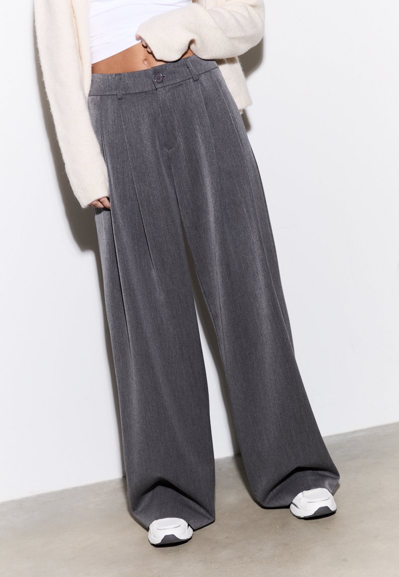 Pantalon gris à jambes larges avec un design plissé, texture moyenne, fermeture à boutons sur le devant, porté avec des baskets blanches.