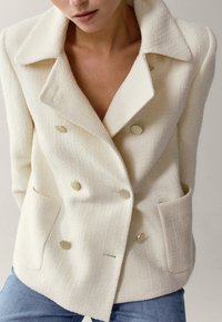 Blazer bianco doppiopetto in tessuto testurizzato con grandi bottoni dorati, dotato di due tasche frontali e un design del colletto strutturato.