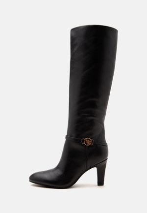 Botte femme en cuir noir montant jusqu'aux genoux avec bout pointu, talon moyen et sangle décorative avec boucle ronde dorée autour de la cheville.
