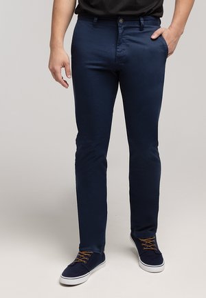 Granatowe, dopasowane bawełniane spodnie z przednimi kieszeniami i zapięciem na guziki. Noszone z ciemnymi sneakersami z brązowymi sznurowadłami.