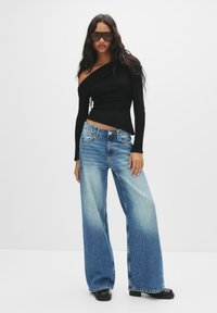 Zwarte off-shoulder top en lichtblauwe wijdvallende jeans met een vervaagde afwerking. Kenmerken een hoge tailleband en flinke zwarte laarzen. Grote zonnebril.