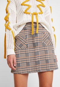 Jersey de punto blanco con acentos amarillos, sobre una mini falda a cuadros en tonos marrón, negro y naranja, que presenta dos bolsillos frontales.