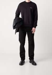 Felpa nera con collo rotondo e logo, abbinata a pantaloni neri e stivali di pelle. Una giacca di pelle nera è appoggiata sul braccio.