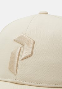 Beige baseball cap lavet af bomuld med en tekstureret overflade, der har en fremtrædende metallic guld broderet detalje i et geometrisk design.