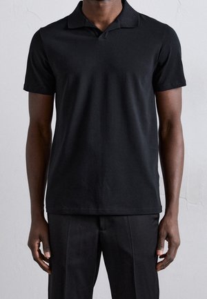 Homme portant un polo noir uni à manches courtes et un pantalon noir, debout contre un mur clair uni.