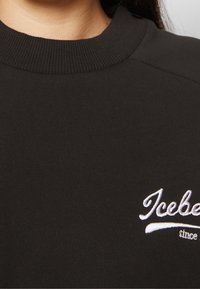 Sweat-shirt noir avec un col côtelé, présentant un texte brodé blanc indiquant "Iceberg since" sur la poitrine. Texture lisse.