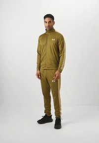 Atletska trenirka olivno zelene barve z jakno z zadrgo in ujemajočimi hlačami, s svetlo rjavimi stranskimi progami ter logotipom na prsih. Črni športni copati.