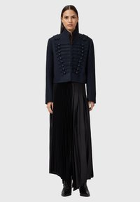 Granatowa kurtka typu cropped z guzikami w stylu wojskowym i naramiennikami, zestawiona z długą, czarną plisowaną spódnicą, która podkreśla gładką fakturę i elegancki design.