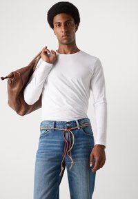 Weißes Langarmhemd, blaue Jeans mit buntem Kordelzug und einer braunen Ledertasche. Glatter Stoff, lockere Passform, lässiger Look.