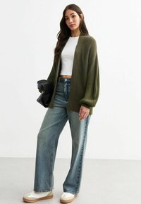 Olive green knitted cardigan with loose sleeves, paired with a white cropped t-shirt and wide-leg light blue jeans. Beige sneakers featured.