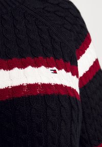 Navy blue strikkegenser med røde og hvite horisontale striper. Har et lite merkelogo på stripen. Teksturert stoff.