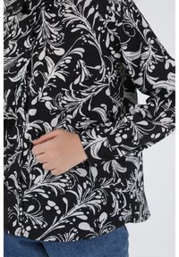 Black button-up shirt with white floral pattern, featuring a relaxed fit, long sleeves, and textured fabric. Hand visible in front pocket area.