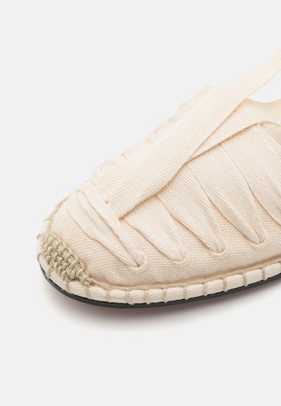 Zapato de textil beige con parte superior tejida, suela plana de goma y detalles trenzados en la punta. Presenta correas de tela cruzando en la parte superior.