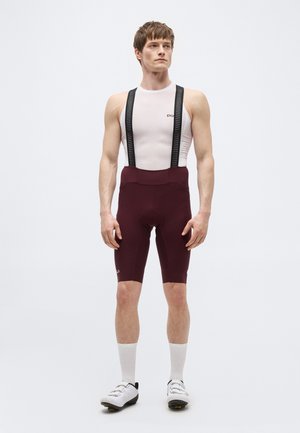 Egy férfi áll, fehér ujjatlan aláöltözetet és bordó kerékpáros kantáros sortot visel fekete pántokkal, fehér kerékpáros cipővel és zoknival.