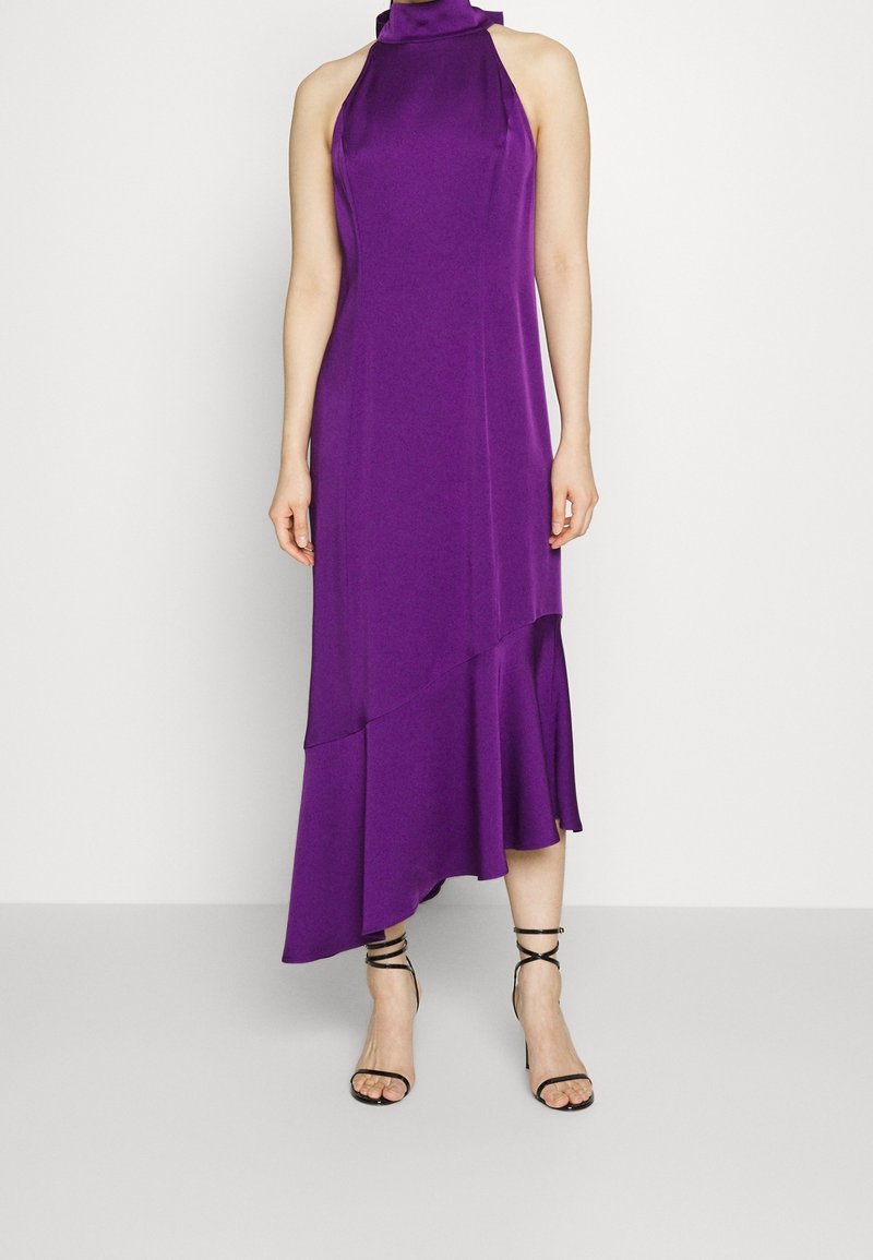 Robe sans manches violette à col halter avec un ourlet asymétrique associée à des sandales à talons noirs à lanières sur une personne devant un fond uni.