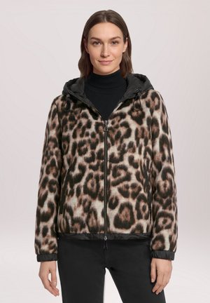 Femme portant une veste à capuche zippée avec un imprimé léopard marron et noir et un pantalon noir, debout devant un fond clair uni.