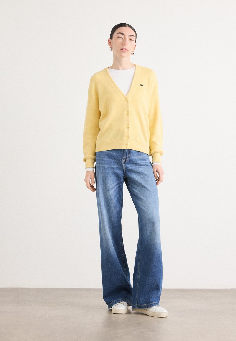 Gelbes Cardigan mit V-Ausschnitt und vorderem Knopfverschluss, kombiniert mit hellblauen weit geschnittenen Jeans und weißen Sneakers. Strukturierter Strickstoff.