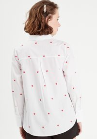 Chemise blanche à manches longues avec broderie en forme de cœur rouge. Comprend un col à boutons, un ourlet arrondi et des manches à poignet. Texture de tissu lisse.