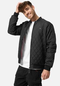 Veste bomber noire matelassée avec fermeture éclair sur le devant, poignets côtelés et poches latérales. Présente un logo sur la poitrine et une texture lisse.