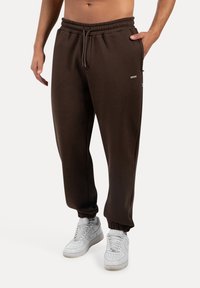 Braune Sweatpants mit konischem Bein, elastischem Bund und Kordelzug. Mit Seitenfächern und einem kleinen Logo-Detail. Einfache Textur.