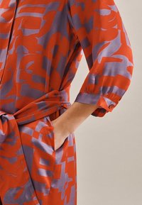 Vestido de patrón rojo y púrpura con mangas de tres cuartos, cintura ceñida y bolsillos laterales; textura sedosa y diseño geométrico audaz.