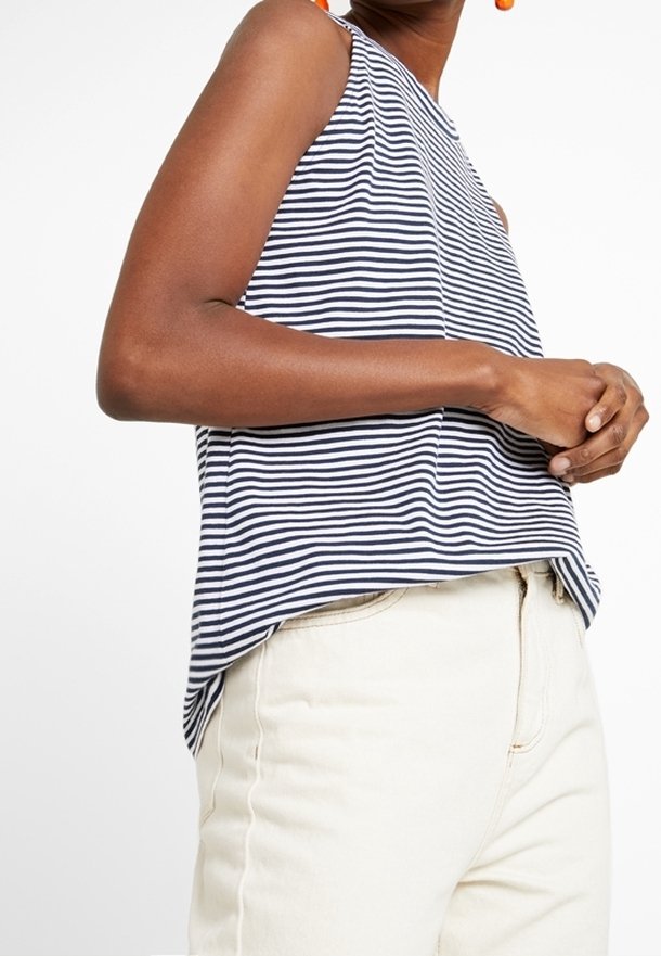 Top senza maniche a righe in navy e bianco, con una vestibilità rilassata, scollatura alta e orlo leggermente curvato. Abbinato a pantaloni di colore chiaro.