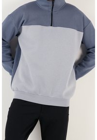 Sudadera de pullover en gris claro y azul con cuello alto, cremallera y paneles en contraste; tejido suave con una textura lisa.