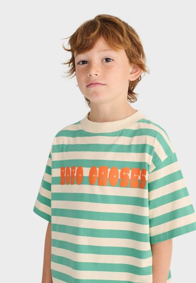 Rothaariges Kind mit grün-weiß gestreiftem T-Shirt und großem orangefarbenem Text "BOBO CHOSES" auf der Vorderseite, neutraler Hintergrund.