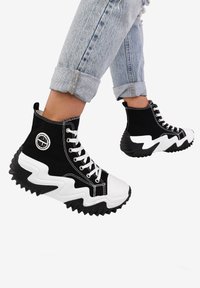 Zapatillas altas de lona negra con punteras de goma blanca y suelas en zigzag. Presentan cordones blancos y un logo en el lateral. Acabado texturizado.