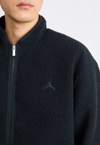Giacca in pile nera con superficie testurizzata, colletto alto e zip completa. Presenta un piccolo logo scuro nella zona sinistra del petto.