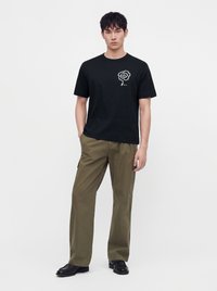 Homme debout portant un t-shirt noir avec un logo en spirale blanche et un pantalon cargo vert olive avec des chaussures noires, sur un fond blanc uni.