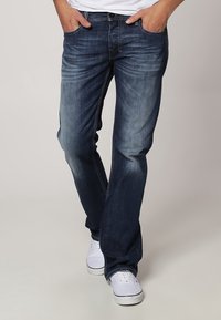 Jeans de mezclilla azul oscuro con corte recto, detalles desgastados, diseño de cinco bolsillos y costuras amarillas en contraste, combinados con zapatillas blancas.