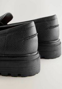 Mocasines de cuero negro con parte superior texturizada, suelas de goma gruesas y detalles de costura elevada. El tacón tiene un diseño robusto y resistente.