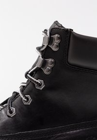 Botte de randonnée en cuir noir avec des lacets métalliques, des œillets hexagonaux et des semelles en caoutchouc noir. Présente une texture lisse et des coutures contrastées.