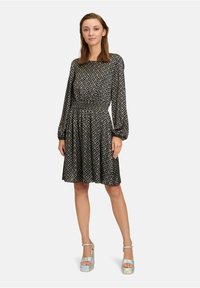 Schwarzes Kleid mit geometrischem weißen Muster, langen Ärmeln, elastischem Bund und ausgestellt Rock. Kombiniert mit glänzenden Plateausandalen.