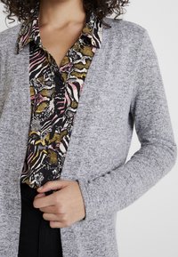 Cardigan gris en tissu texturé, superposé sur une chemise à motifs avec des dessins noirs, roses et jaunes, comportant une fermeture à boutons.