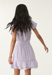 Abito corto in lavanda con maniche a campana, vita arricciata, orlo con volant e una leggera texture. La vista dal retro mostra i capelli che scendono sulle spalle.