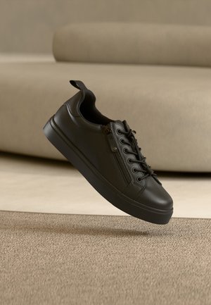 Sneaker in pelle nera con lacci e cerniera laterale, sospesa sopra un tappeto beige in un ambiente interno dai toni neutri.