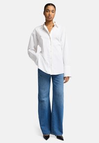 Camicia bianca con colletto a punta, abbinata a jeans a gamba larga blu chiaro e tacchi neri a punta, che mostrano un'eleganza informale.