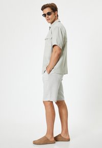 Chemise à manches courtes gris clair avec deux poches poitrine, associée à un short gris clair et des mocassins marron. Le modèle porte des lunettes de soleil noires.