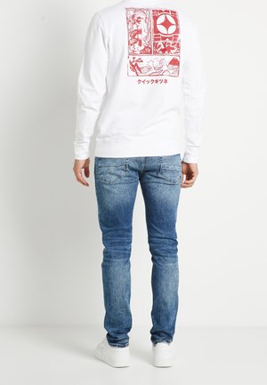 Mężczyzna w białej koszuli z długim rękawem, z czerwonym nadrukiem i japońskim napisem na plecach, ubrany w niebieskie dżinsy i białe trampki, stoi odwrócony tyłem.