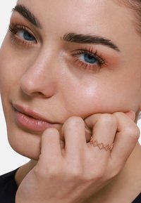 Rosenvergoldeter Ring mit einem wellenförmigen Design, ausgestattet mit einem zarten Kettenmuster. Der Hautton ist hell, mit einer glatten Textur und minimalem Make-up.