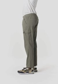 Persona con pantalones cargo verde oliva con bolsillos de parches y zapatillas beige, negras y blancas, de pie contra un fondo claro y liso.
