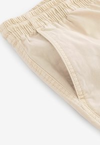 Shorts beiges avec une taille élastique, texture lisse, poche latérale, finition avec des coutures soignées. Le tissu semble léger et respirant.
