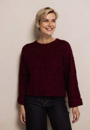 Smiling woman with short blonde hair wearing a textured dark red sweater and dark pants, standing against a plain light background.