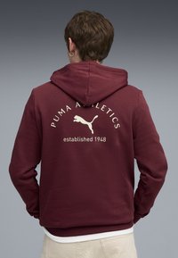 Bordeaux hættejakke med kengurulomme, justerbar hætte og hvid tekst med "PUMA ATHLETICS" samt et logo på ryggen.