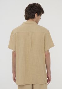 Camicetta a maniche corte di colore beige chiaro, realizzata in tessuto testurizzato. Presenta un colletto e una piega centrale sul retro per una maggiore struttura.